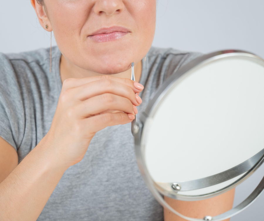 Vrouw kijkt in de spiegel en trekt een haar uit haar kin met een pincet, een teken van hormonale disbalans en overbeharing
